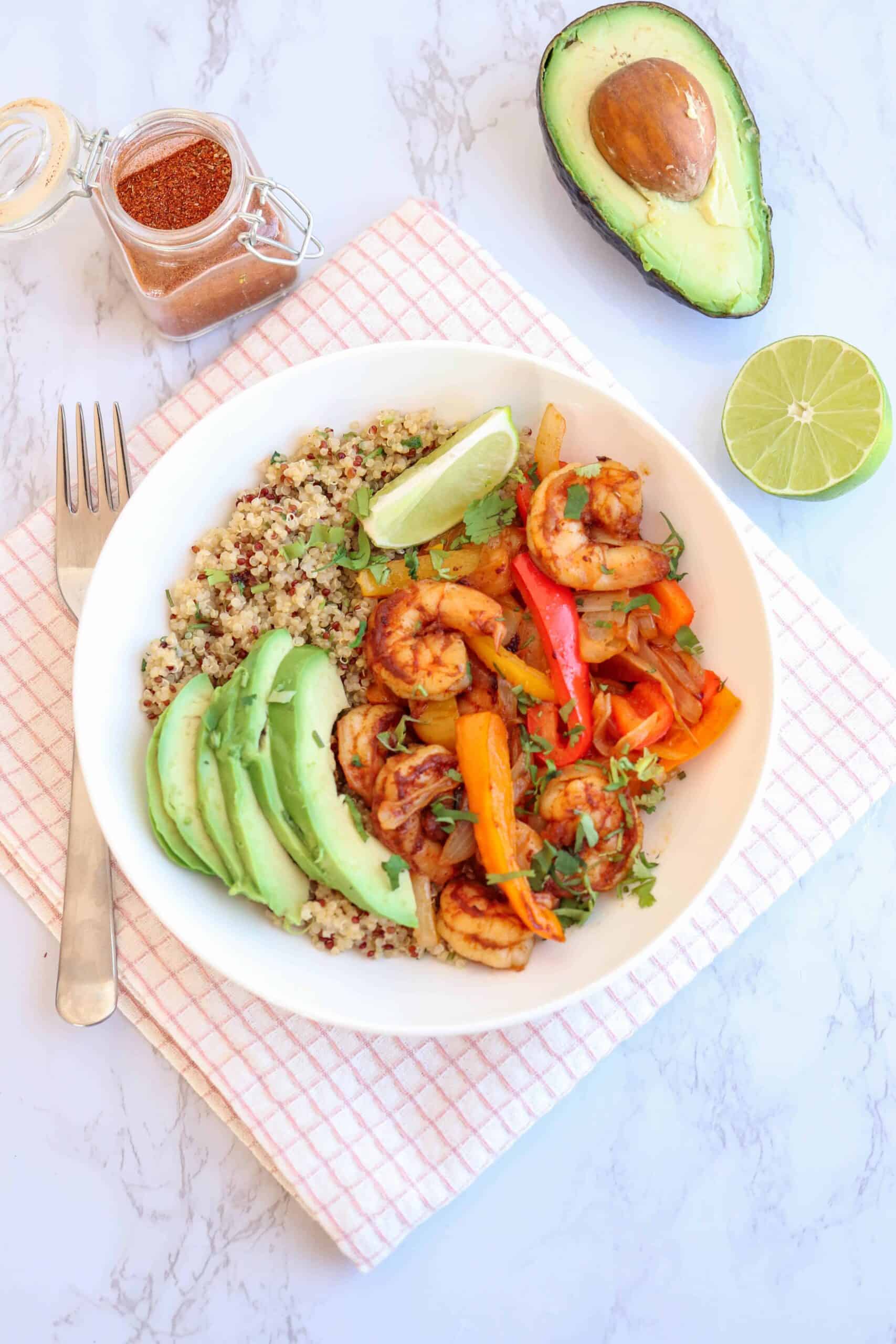 a bowl of shrimp fajita with avocado over quinoa on white background with half a lime and avocado