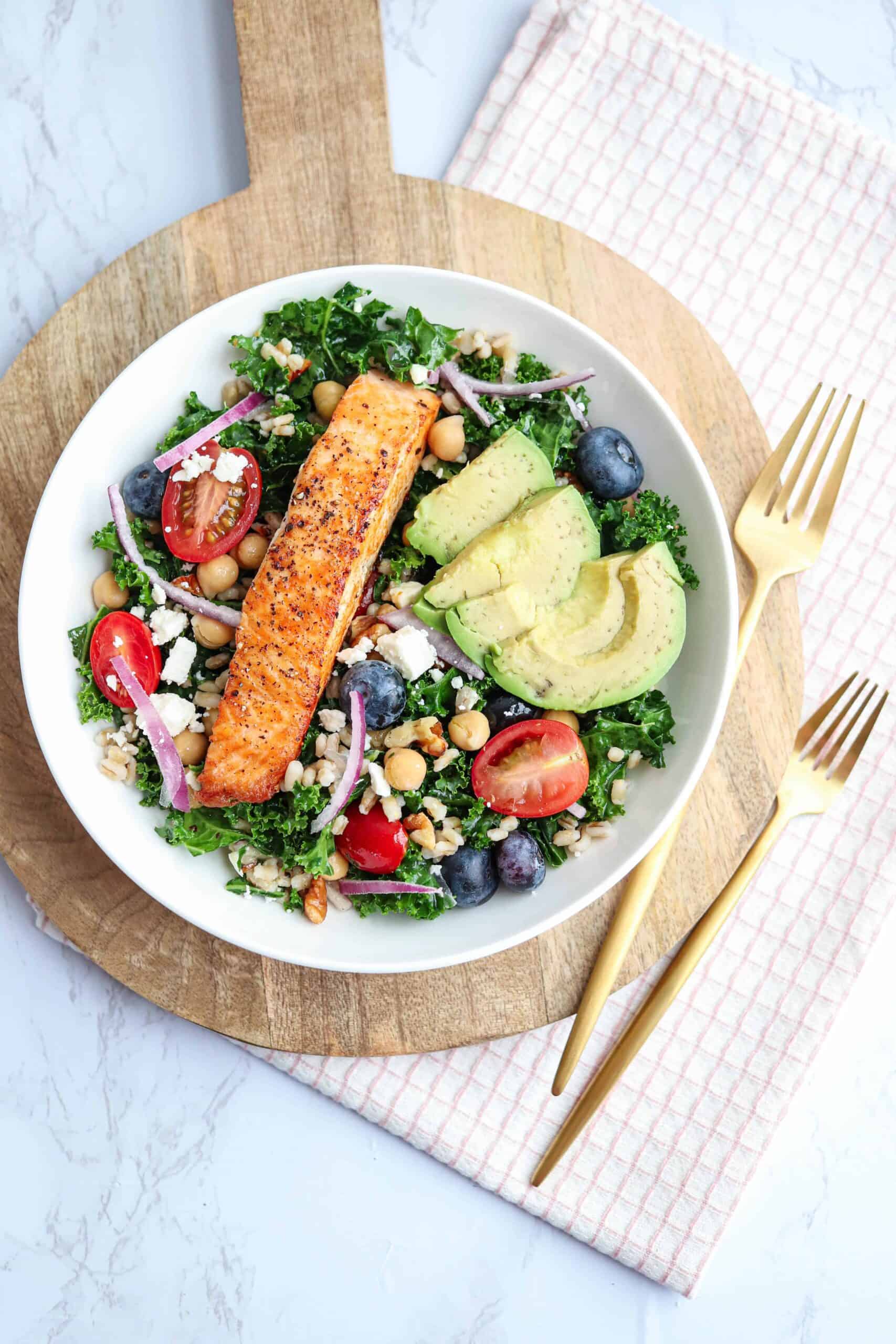 a bowl of kale barley salad bowl with seared salmon and avocado on a wooden board on marble counter