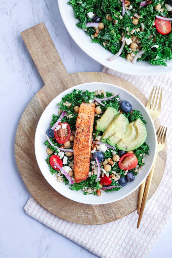 a bowl with kale and barley salad and salmon on a wooden board with 2 gold forks