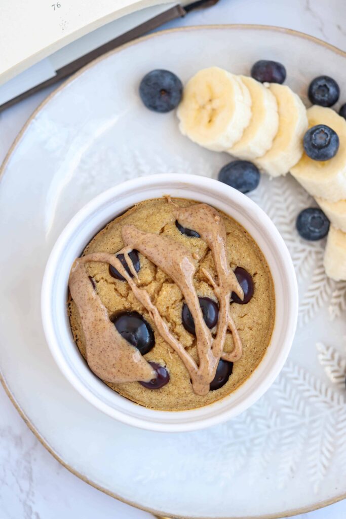 white ramekin with banana baked oats with blueberries and almond butter on top on a plate with a side of bananas and blueberries