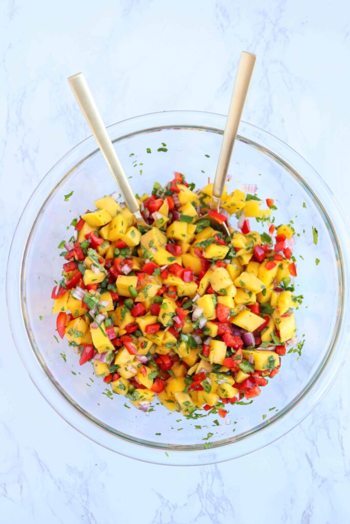 a glass bowl with fresh mango salsa and 2 gold spoons on white marble surface