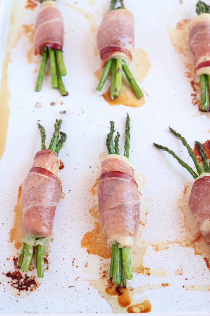 prosciutto wrapped asparagus stuffed chicken baked on a white baking sheet.