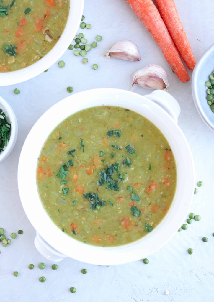 a white bowl of split pea and ham soup garnished with parsley on gray countertop with carrots, garlic cloves and green peas around