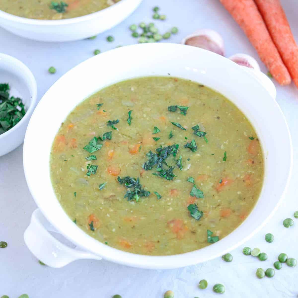 a white bowl with split pea soup with carrots and parsley leaves on gray surface with carrots, split peas and garlic cloves around