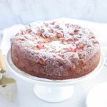 rhubarb sponge cake dusted with powdered sugar on a white cake stand with white marble background