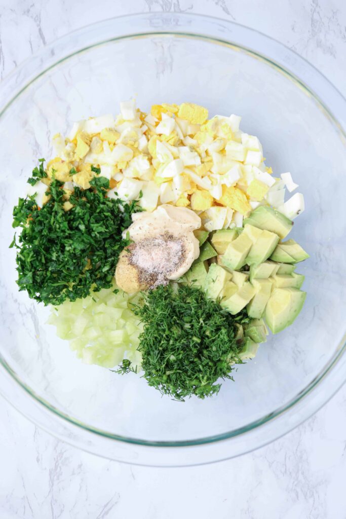 a glass bowl with chopped eggs, avocado, celery, mayo, dill, parsley, mustard, salt and pepper on white marble surface