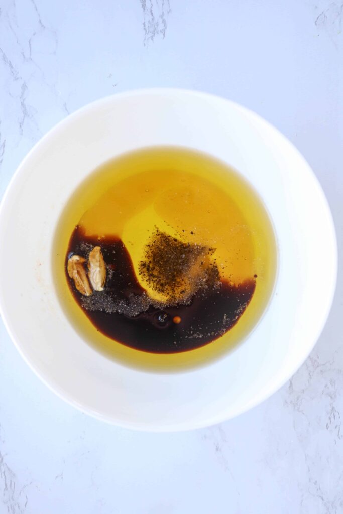 a small white bowl with olive oil, balsamic vinegar, mustard, salt and pepper before mixing on white marble surface