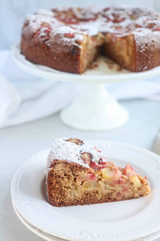 a slice of rhubarb cake dusted with powdered sugar on a white plate, in the background there is the whole cake on a white cake stand with once slice missing