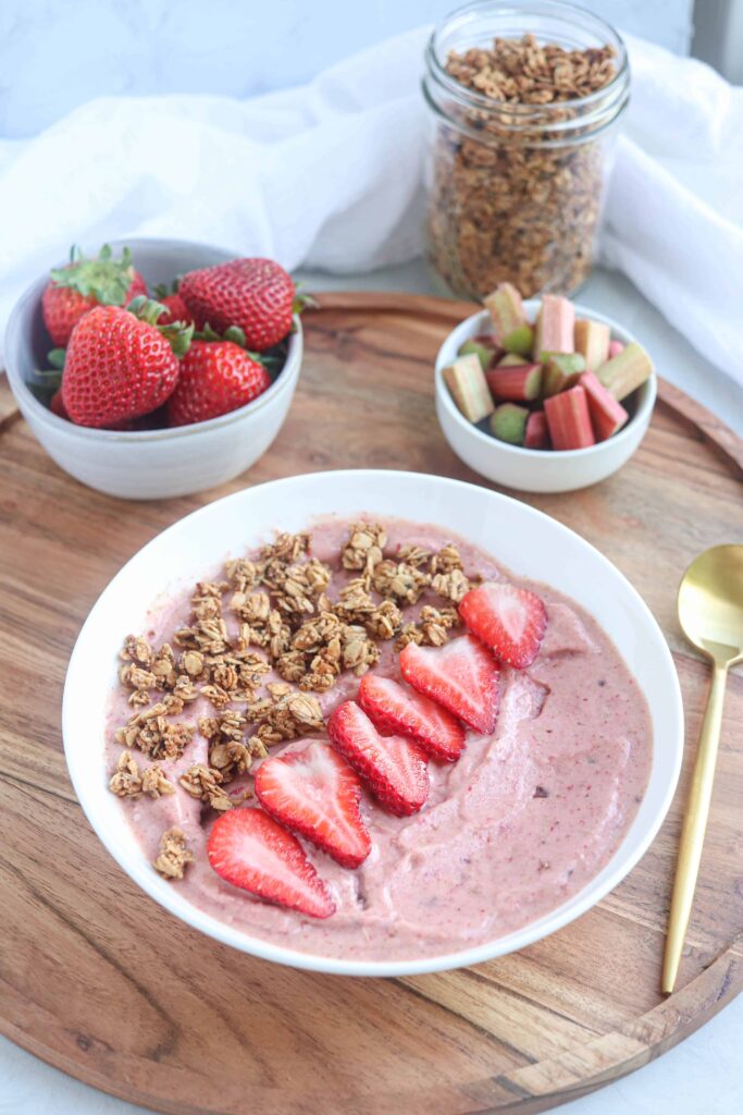 a white bowl with strawberry rhubarb smoothie bowl topped with granola and fresh strawberry slices on a round wooden board. In the background there are dishes with strawberries, rhubarb and granola and a golden spoon