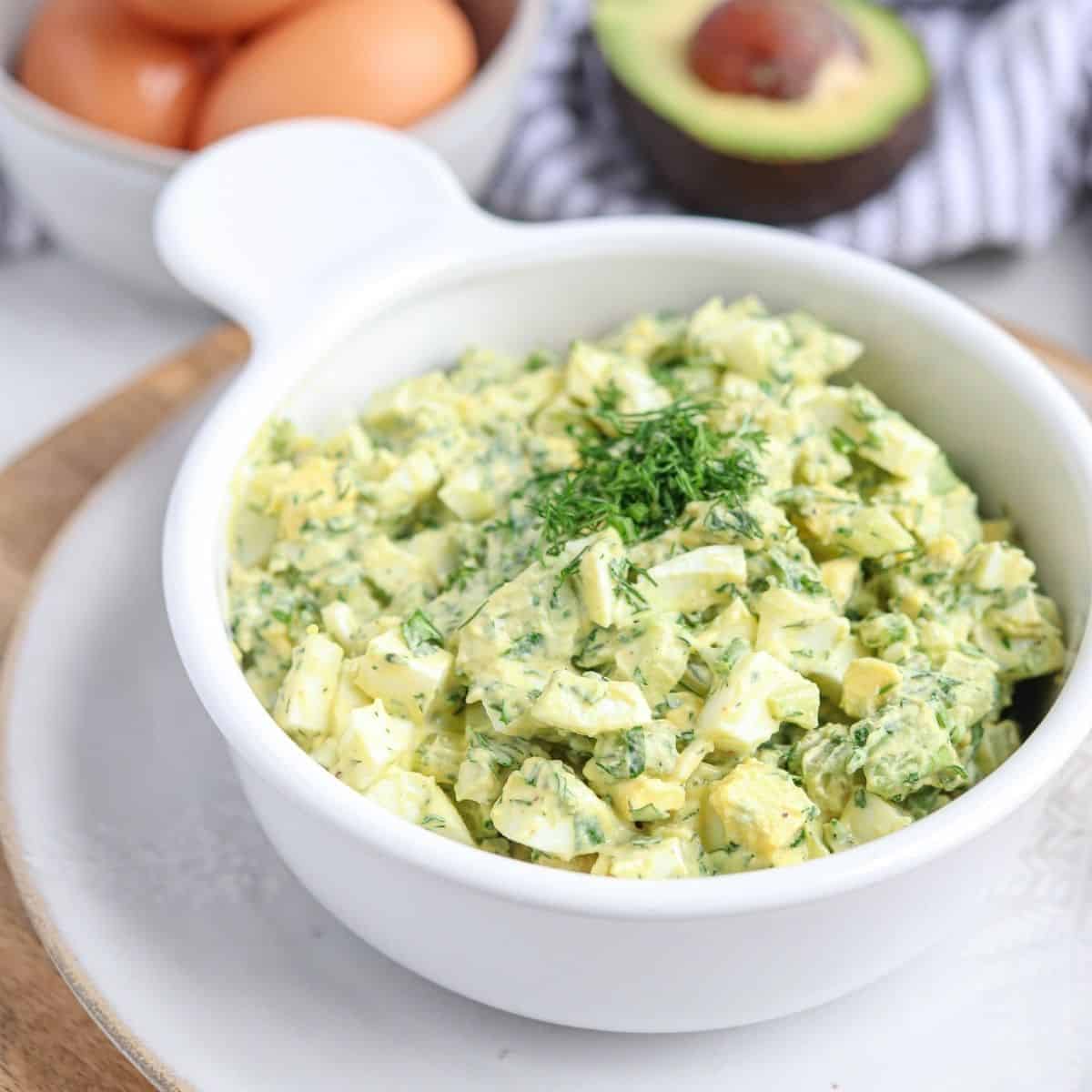 egg avocado salad in a white bowl on a light colored plate with eggs and avocado in the background