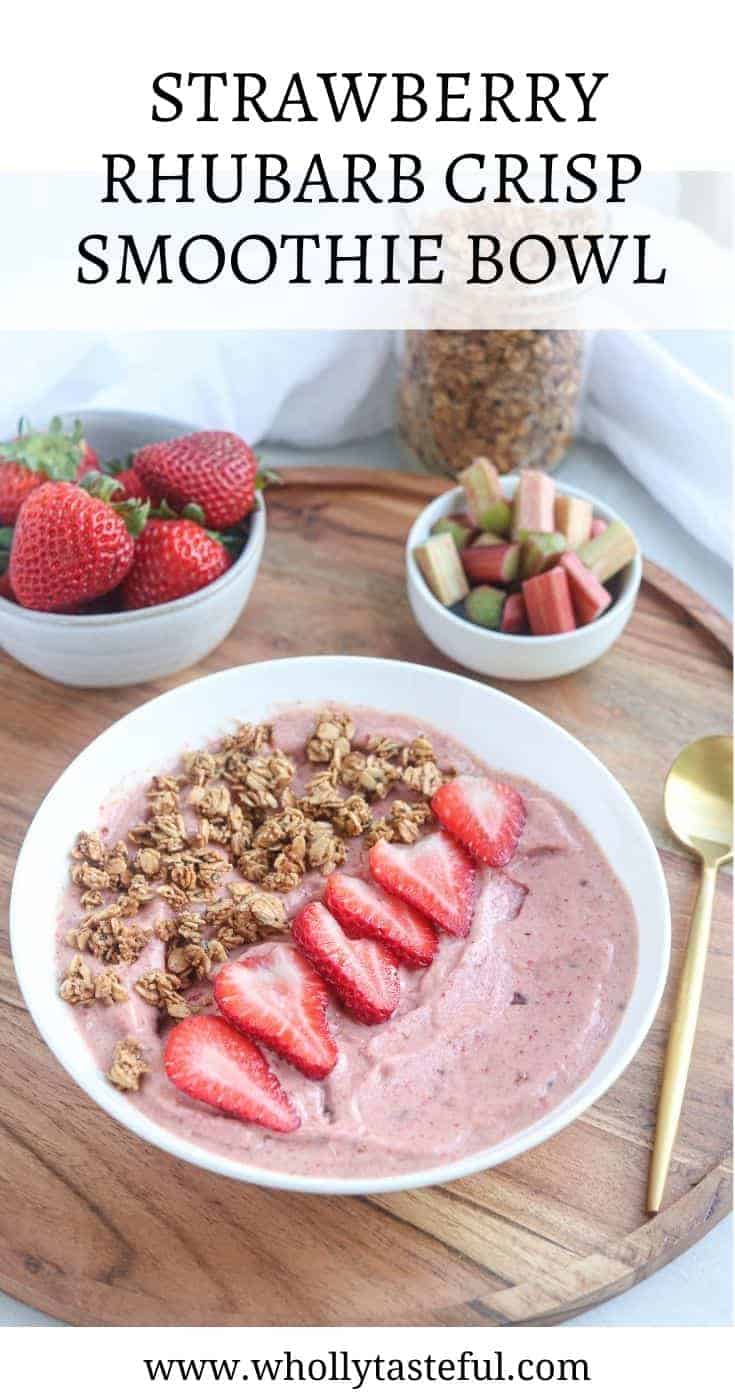 a white bowl with strawberry rhubarb smoothie bowl topped with granola and fresh strawberry slices on a round wooden board. In the background there are dishes with strawberries, rhubarb and granola and a golden spoon with title on top saying "Strawberry Rhubarb Crisp Smoothie Bowl"