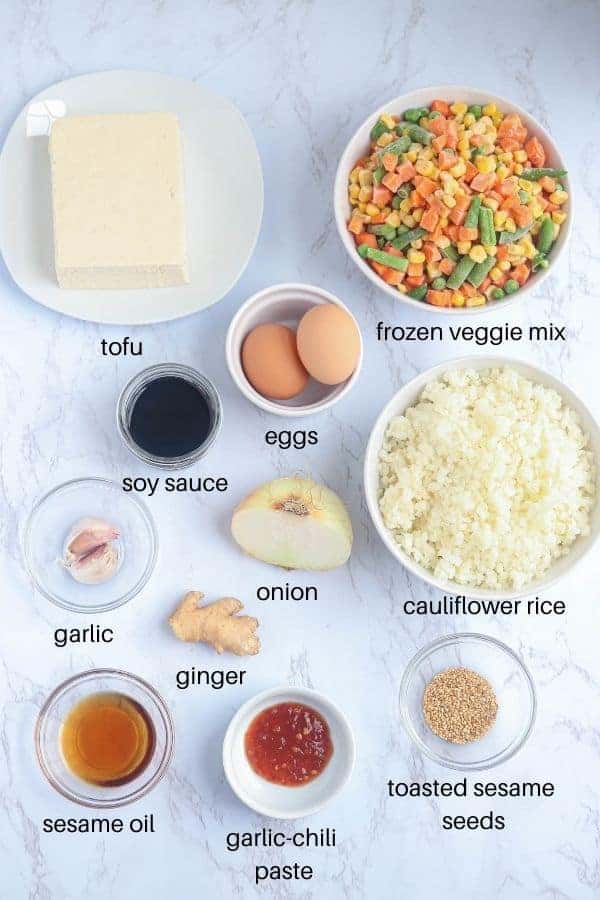 ingredients for making tofu cauliflower fried rice on white marble surface with captions.