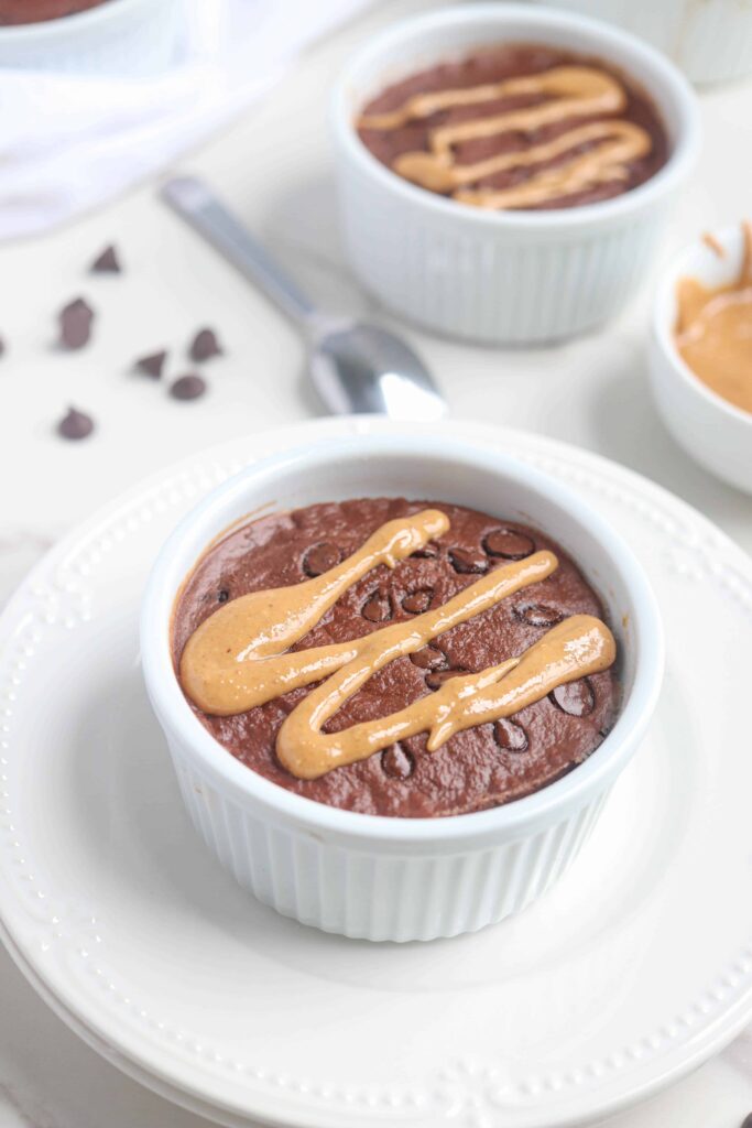chocolate baked oats in a white ramekin on a white plate with chocolate chips and drizzled with peanut butter