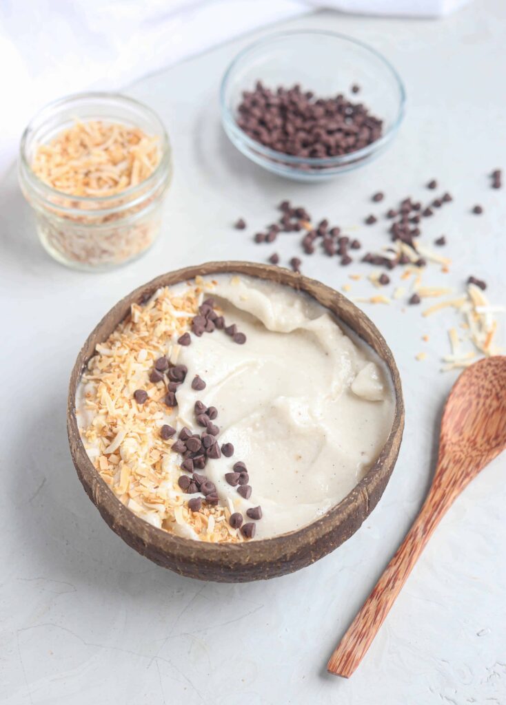 coconut shell bowl filled with coconut vanilla smoothie, chocolate chips, and toasted coconut alongside a wooden spoon.