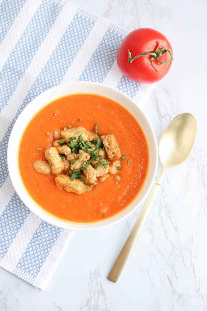 a white bowl of creamy tomato bean soup with croutons and chopped herbs on top on a blue and white striped kitchen towel.