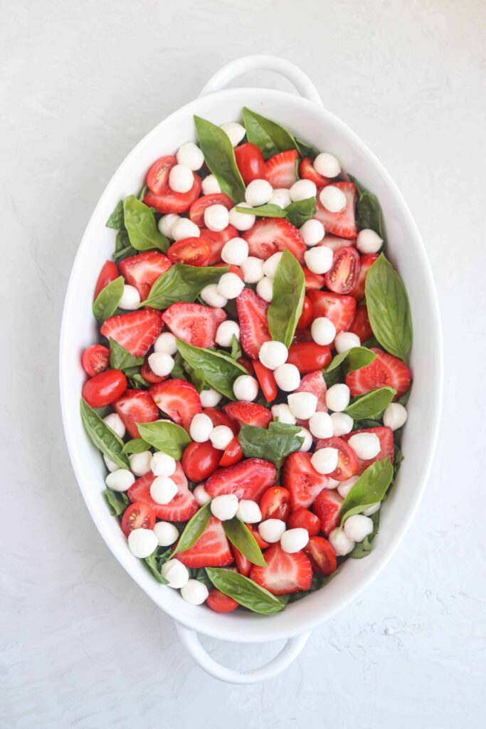 an oval dish with chopped baby spinach, strawberries and cherry tomatoes, mozzarella balls and basil leaves