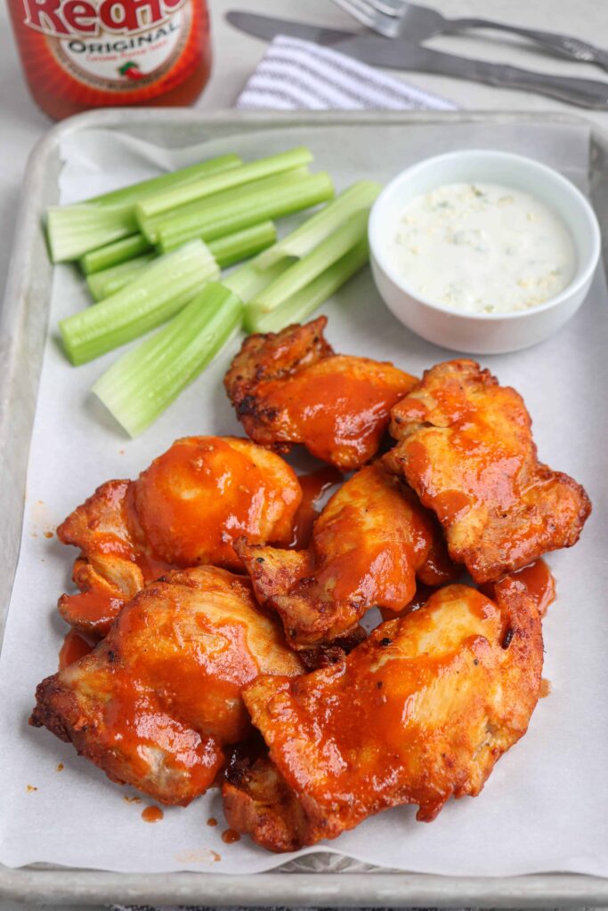 air fryer buffalo chicken thighs on a baking tray served on white paper with a side of celery sticks and blue cheese dressing