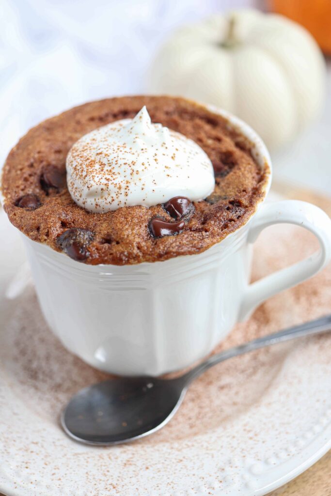 pumpkin mug cake with a dollop of whipped cream and cinnamon in a white teacup on a saucer