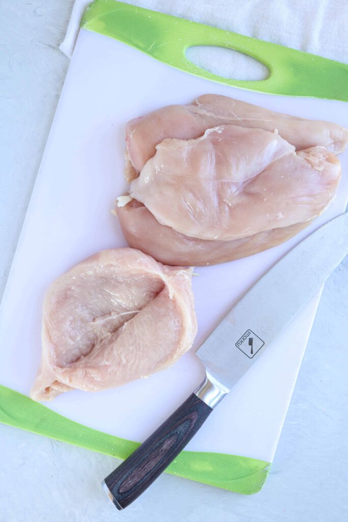 chicken breasts being cut into thin wide slices on a plastic white board. 