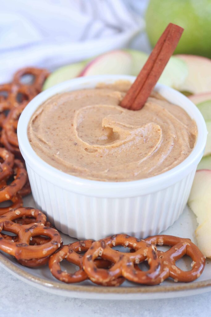 a white ramekin with healthy caramel yogurt dip surrounded by pretzels and apple slices