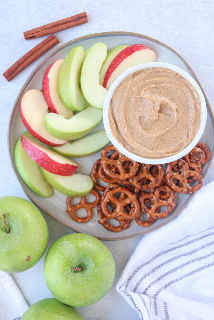 a white ramekin with healthy caramel yogurt dip surrounded by pretzels and apple slices