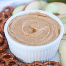A small white ramekin filled with creamy Greek yogurt apple dip, surrounded by sliced apples and pretzels on a plate.