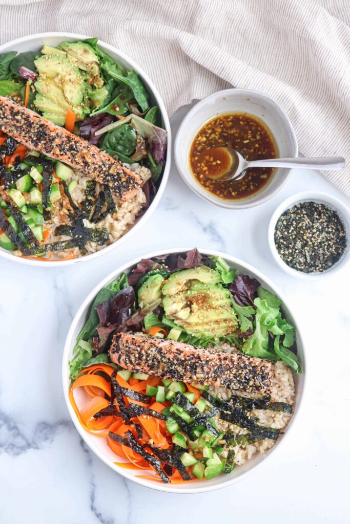 a white bowl with furikake salmon on the bed of greens, avocado, carrots, brown rice and nori on white marble surface