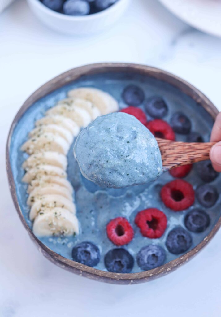 blue spirulina smoothie bowl in a coconut bowl topped with slices bananas, blueberries and raspberries, close up of a bite