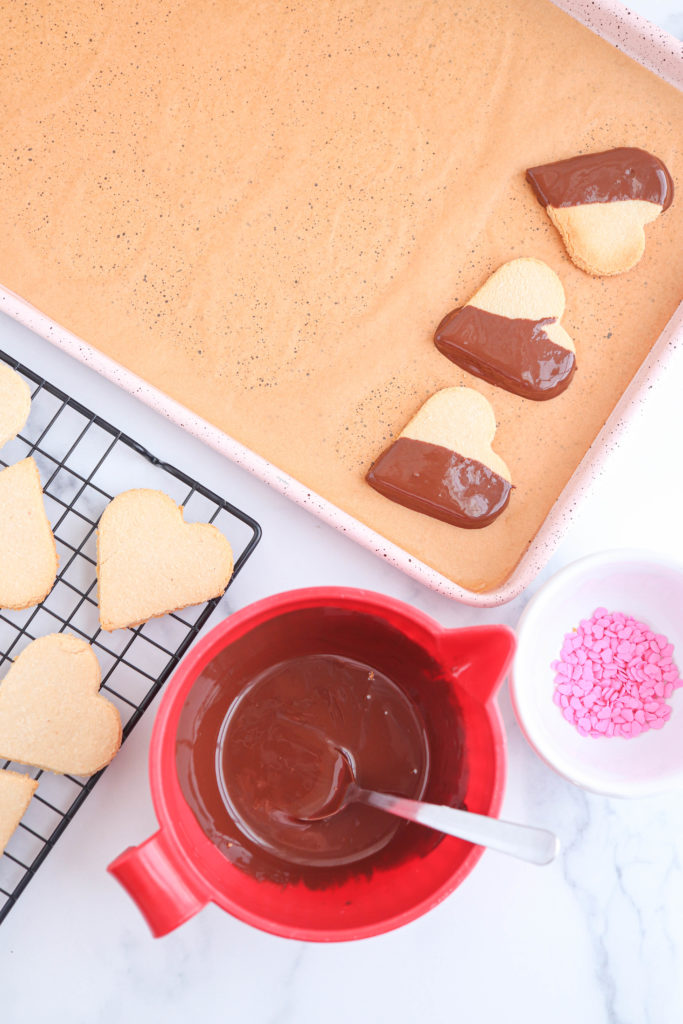 chocolate melted in a red bowl and chocolate dipped heart cookies on a cookie sheet.