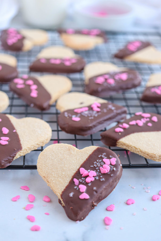heart shaped chocolate dipped vegan shortbread cookies with pink heart sprinkles on a black cooling rack
