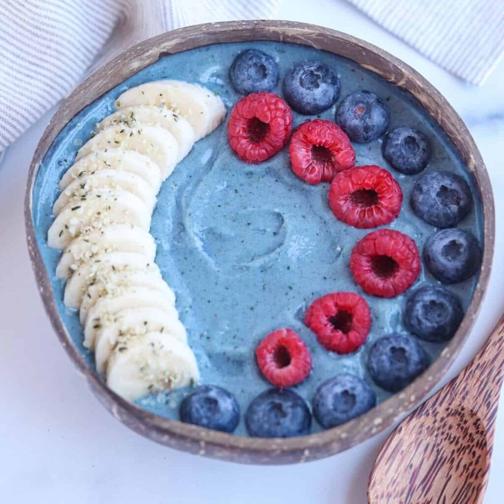 blue spirulina smoothie bowl in a coconut bowl topped with slices bananas, blueberries and raspberries