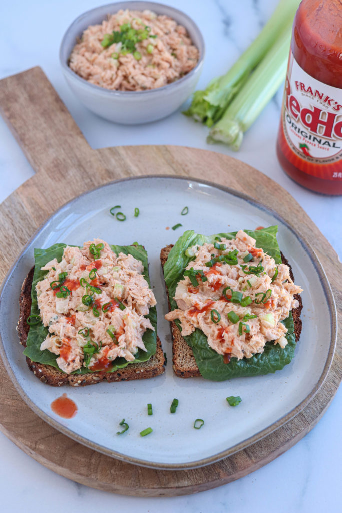 two open faced sandwiches on multigrain bread with lettuce and Buffalo tuna salad drizzled with hot sauce and sprinkled with chopped chives on a gray plate.
