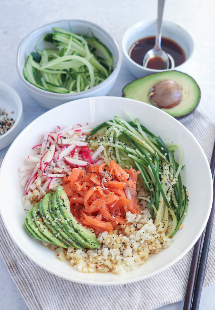 sushi bowl with cauliflower rice, smoked salmon, julienned cucumbers, radishes, sliced avocado and sprinkled with everything bagel seasoning in a white bowl with some ingredients scattered around.