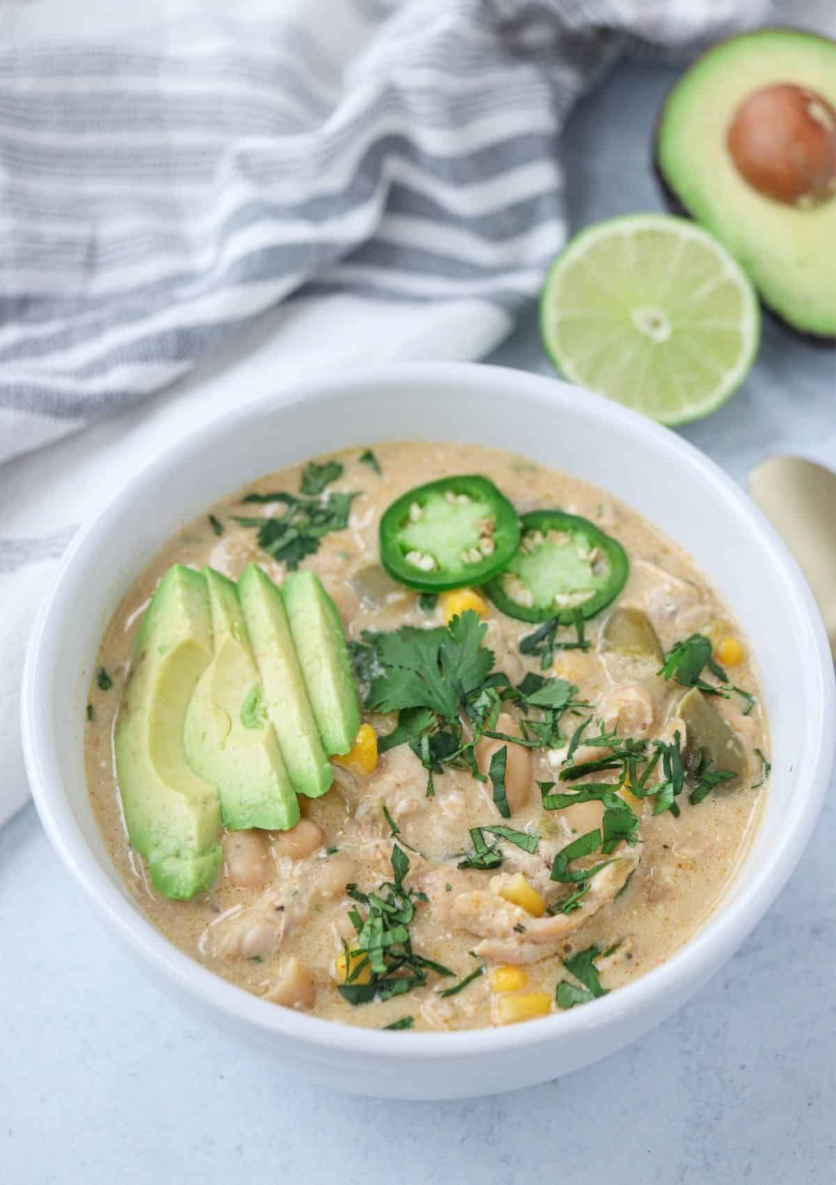 green chicken chili in a white bowl topped with avocado slices, jalapeno peppers and chopped cilantro.