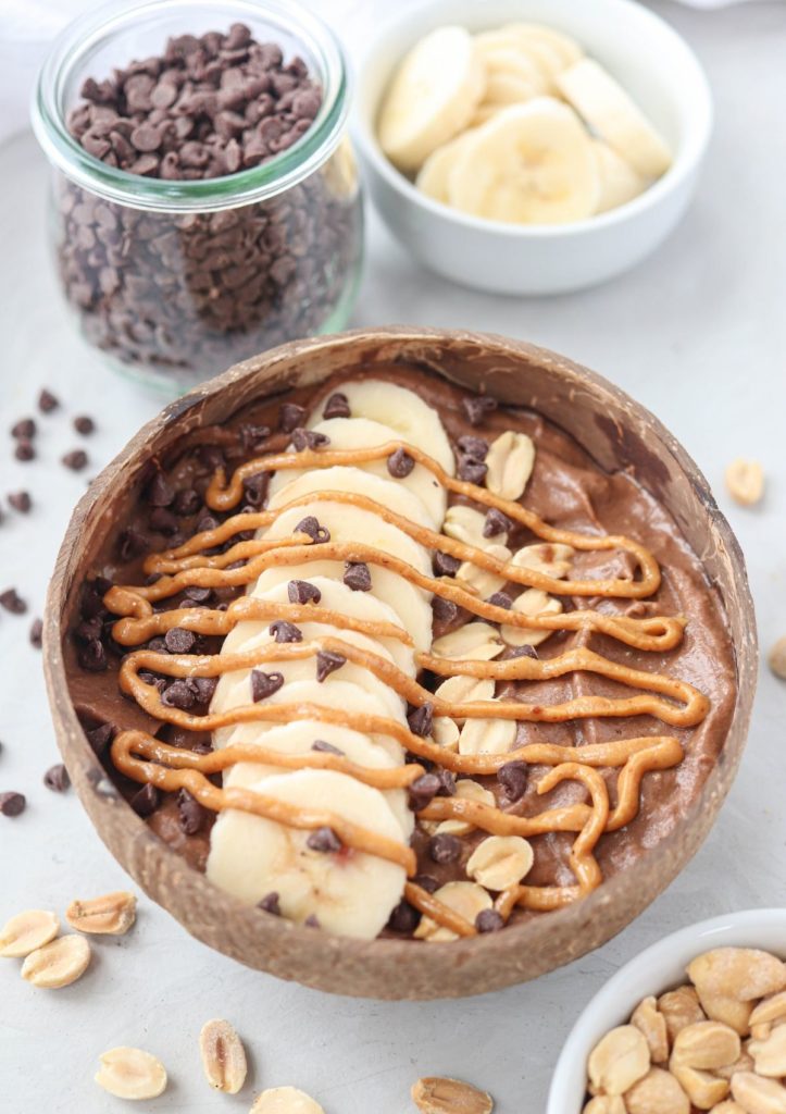 snickers smoothie bowl in a coconut shell bowl topped with sliced bananas, peanuts, chocolate chips and peanut butter.