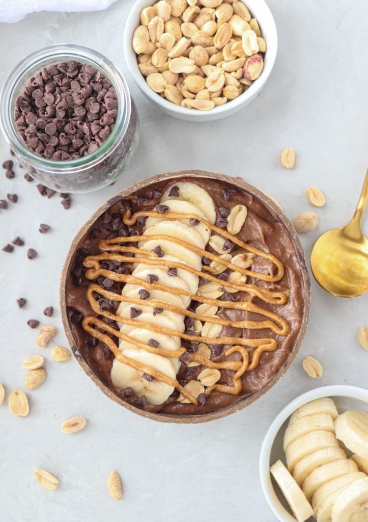 snickers smoothie bowl in a coconut shell topped with banana slices, peanut butter drizzle, peanuts and chocolate chips.