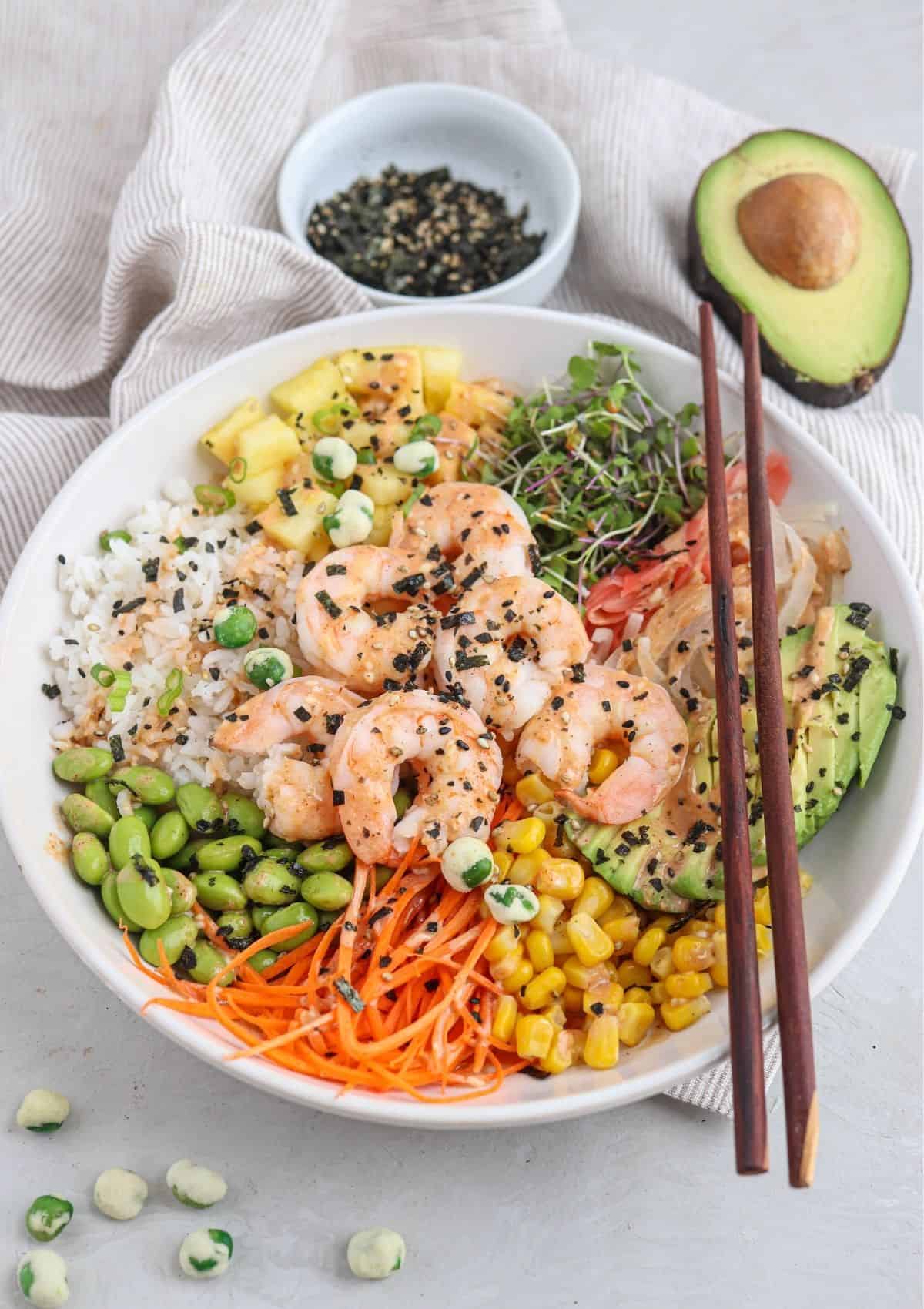 shrimp poke bowl in a white bowl with rice, pineapple, cooked shrimp, edamame, corn, carrots, microgreens and creamy spicy mayo.