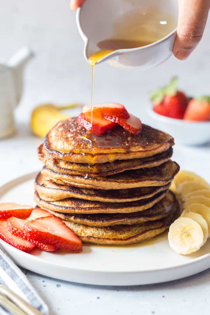 A stack of pancakes topped with sliced strawberries is being drizzled with syrup from a small pitcher. Sliced bananas and more strawberries surround the plate, making this dish a delicious option for healthy banana breakfast recipes.