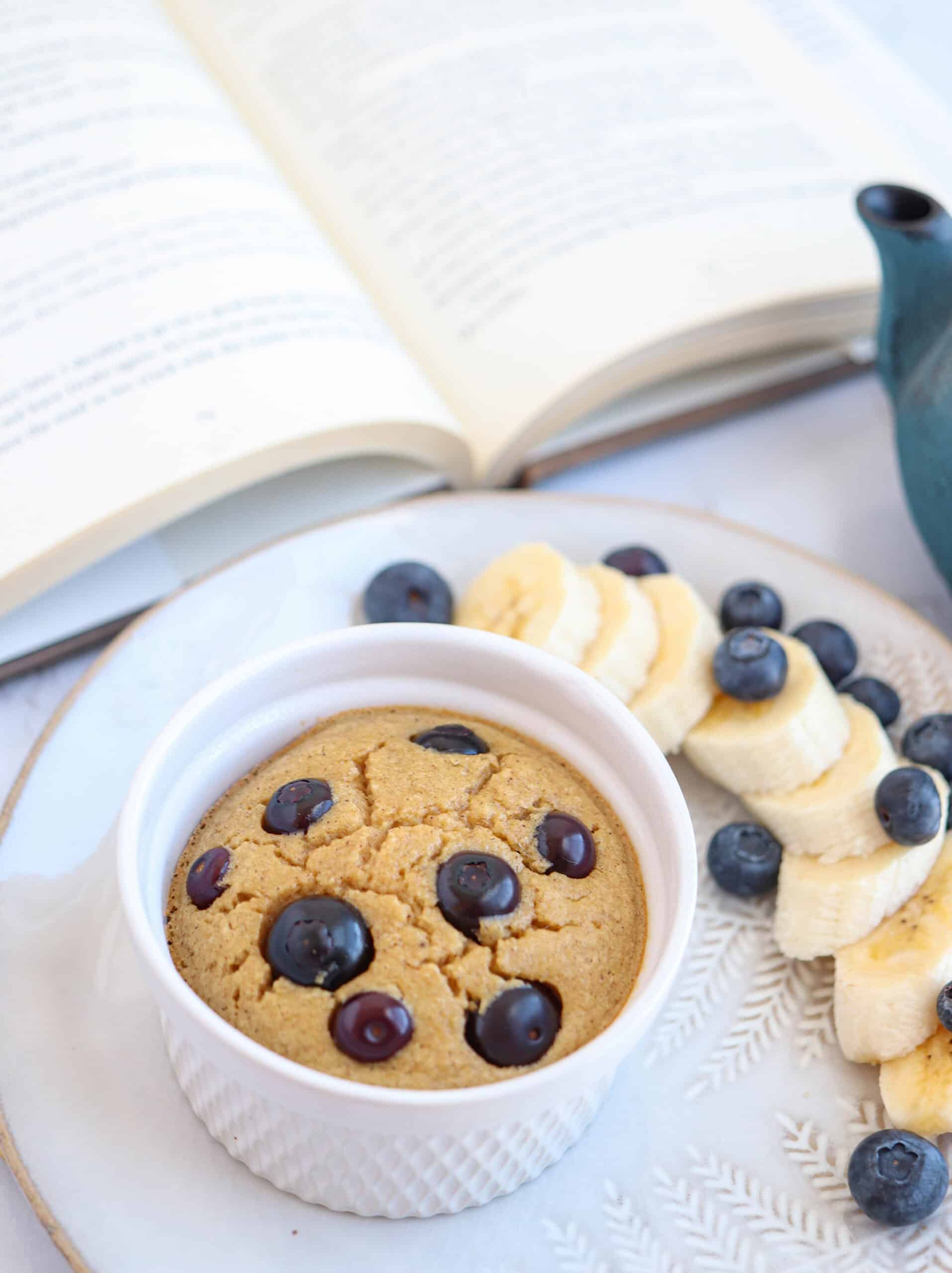 A ramekin with a baked treat topped with blueberries sits on a plate with banana slices and blueberriesโan inviting choice for those seeking healthy banana breakfast recipes. An open book and a blue teapot complete the cozy scene.