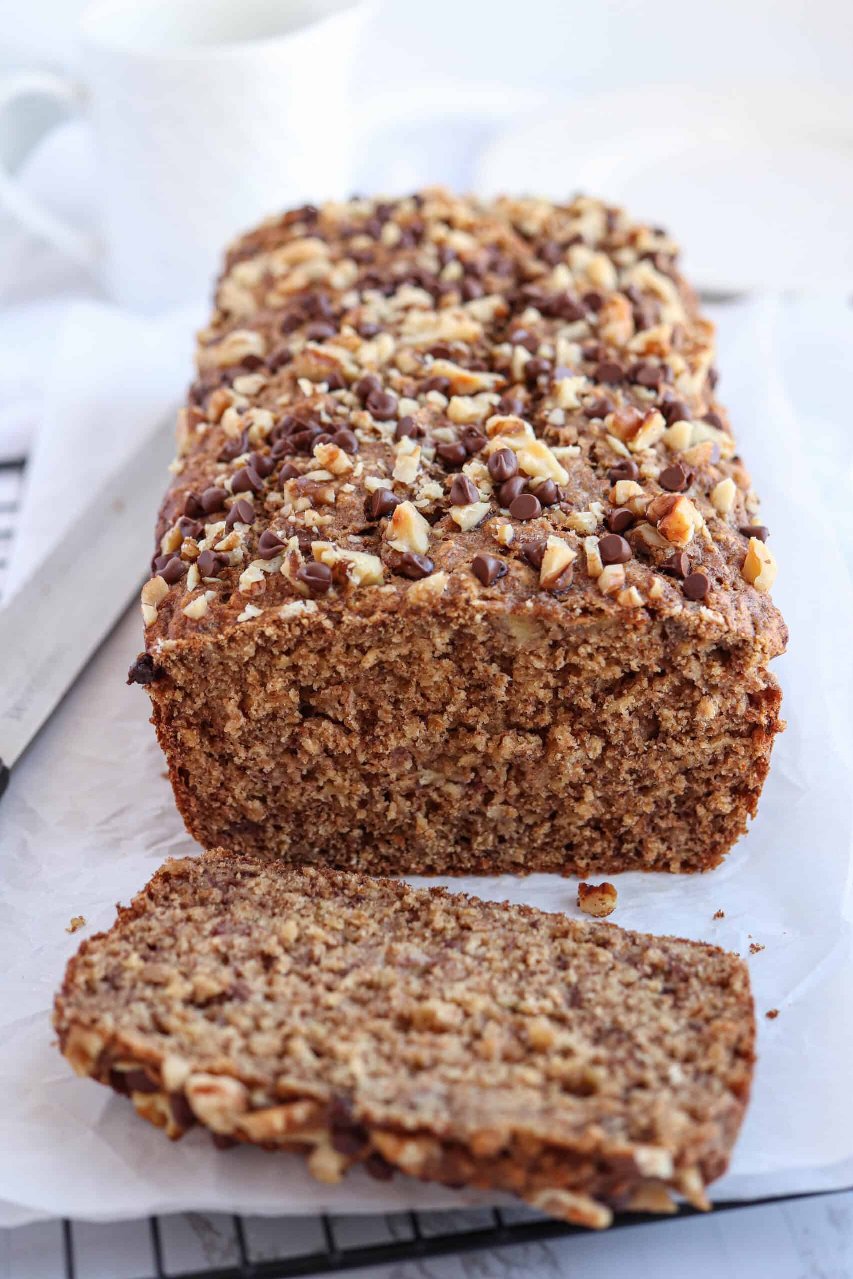 A loaf of banana bread, perfect for healthy banana breakfast recipes, is topped with chopped nuts and chocolate chips on parchment paper. One slice is cut and placed in front, with a knife and white cup in the background.