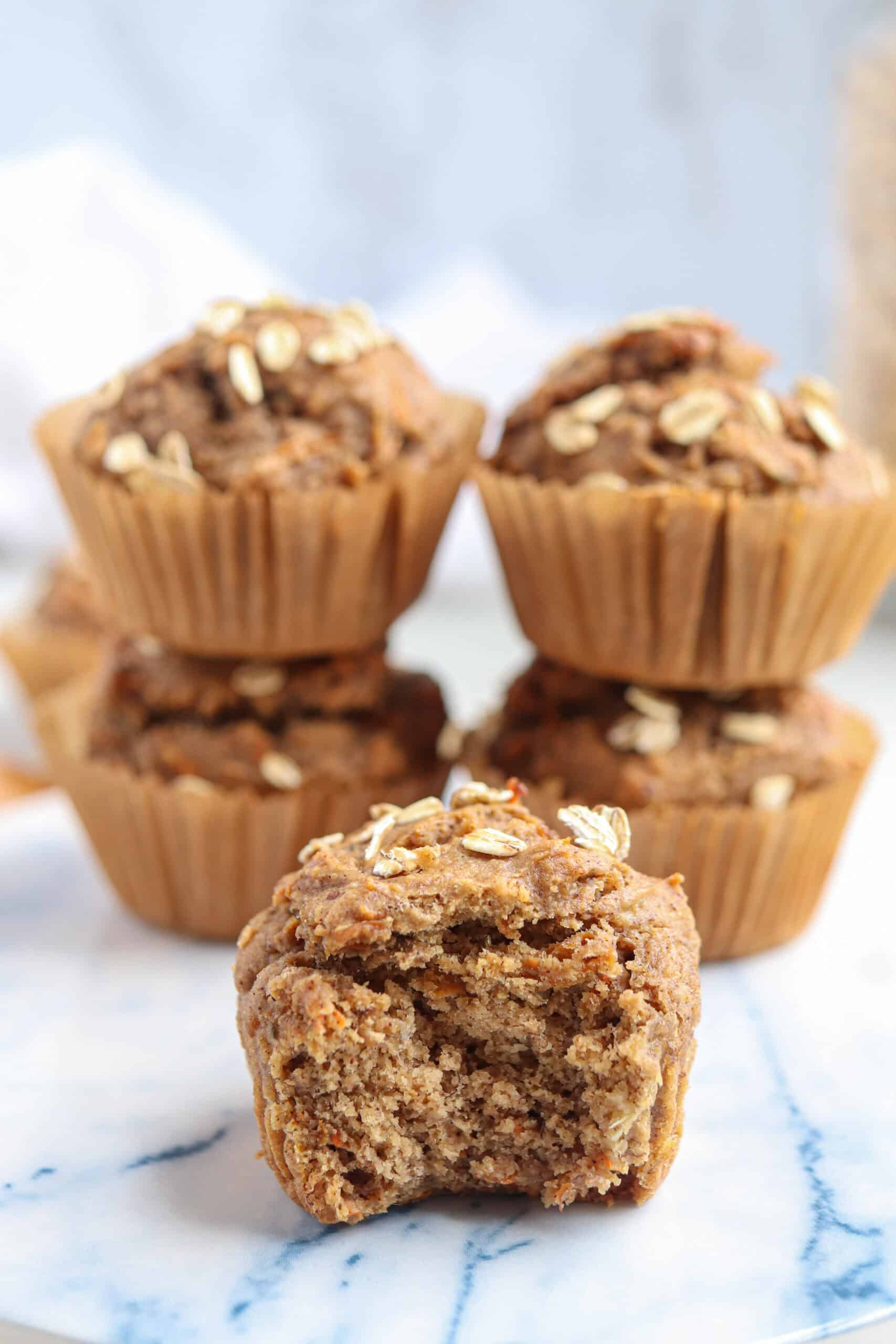 Four oatmeal muffins stacked on a marble surface, with one muffin in front showing a bite taken out. These golden brown treats topped with oats make the perfect addition to your healthy banana breakfast recipes, set against a soft, light background.