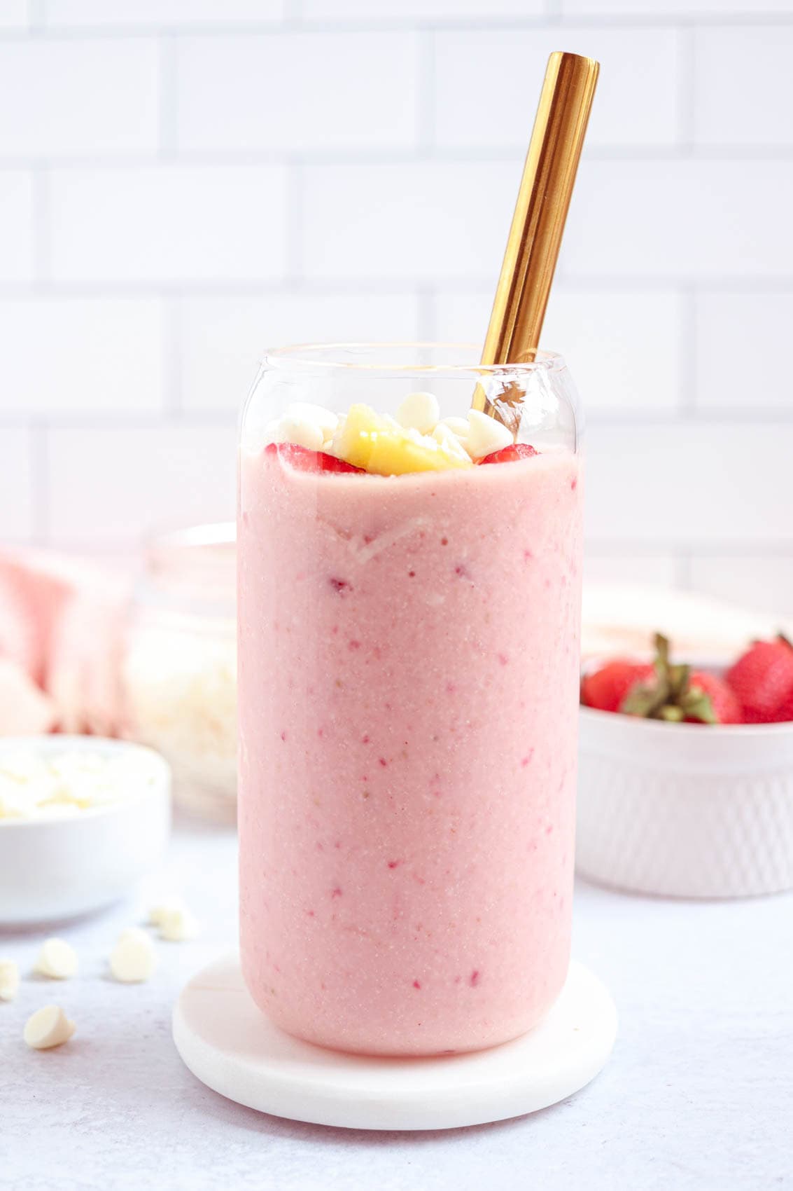 A glass of pink smoothie, inspired by healthy banana breakfast recipes, topped with pineapple chunks, sliced strawberries, and white chocolate chips with a gold straw. The glass sits on a white coaster, with strawberries and a jar in the background.