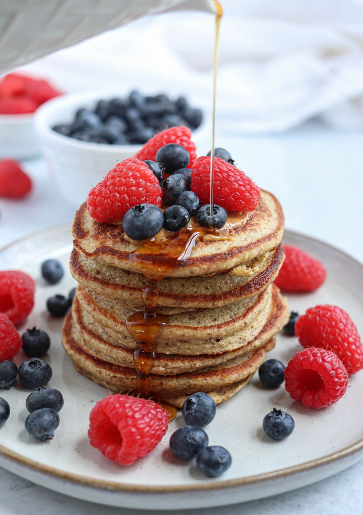 A stack of pancakes topped with fresh raspberries and blueberries, with syrup being poured over themโan irresistible choice for fans of healthy banana breakfast recipes. Extra berries are scattered on the plate, with a bowl of blueberries in the background.