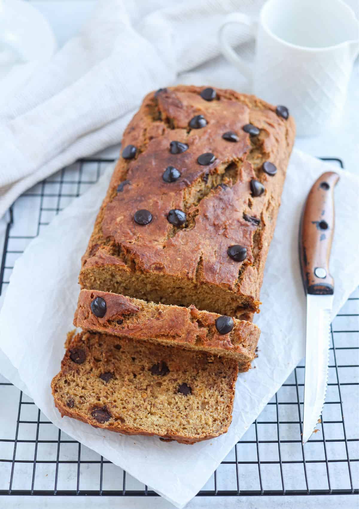 A loaf of chocolate chip banana bread sits on parchment paper atop a cooling rack, with two slices cut. Perfect for those seeking healthy banana breakfast recipes, a knife with a wooden handle lies beside the loaf, and a white mug is in the background.