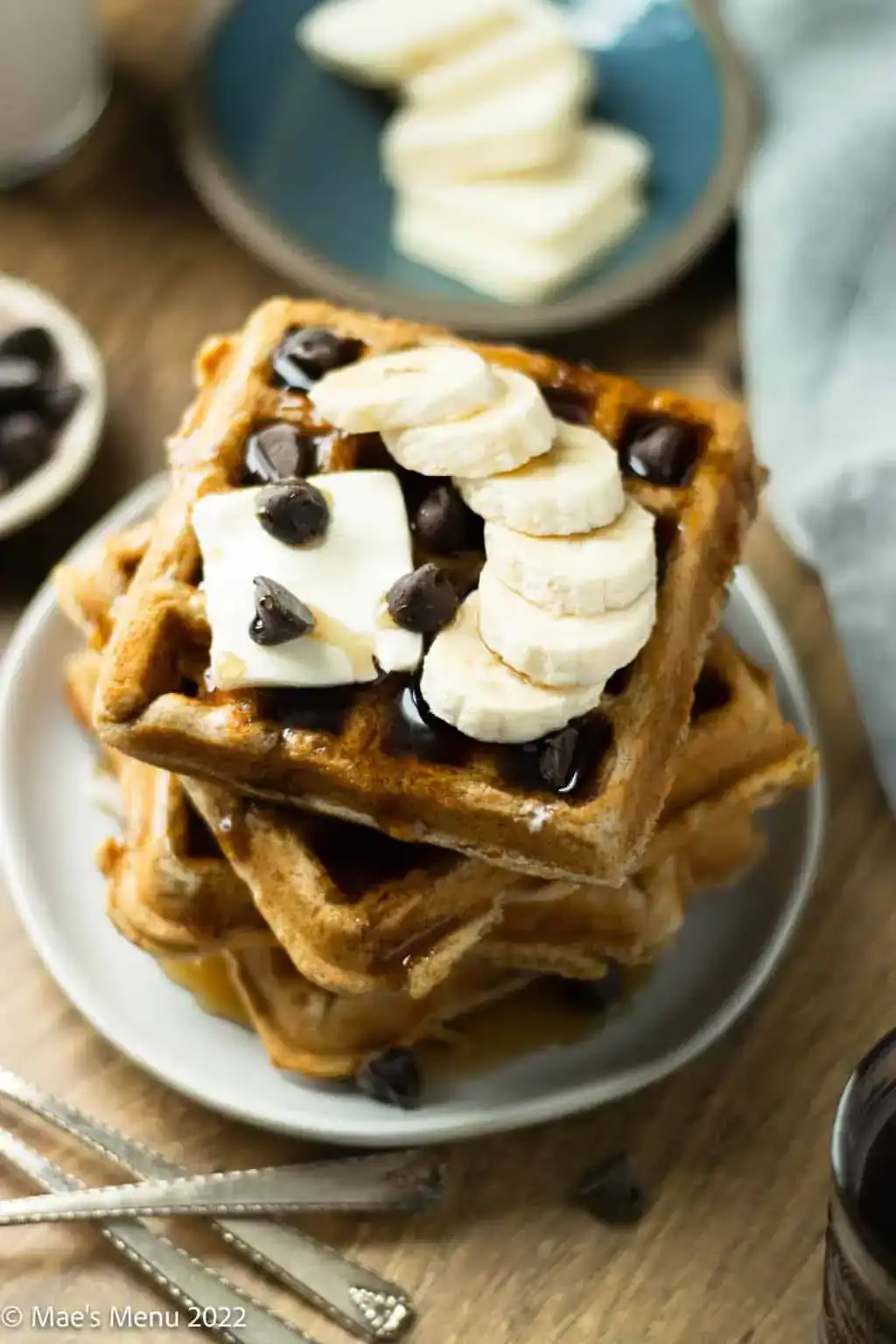A stack of waffles topped with banana slices, chocolate chips, and a pat of butter sits on a plate. Perfect for those seeking healthy banana breakfast recipes, syrup drips down the sides beside a dish of bananas and a cup in the background.