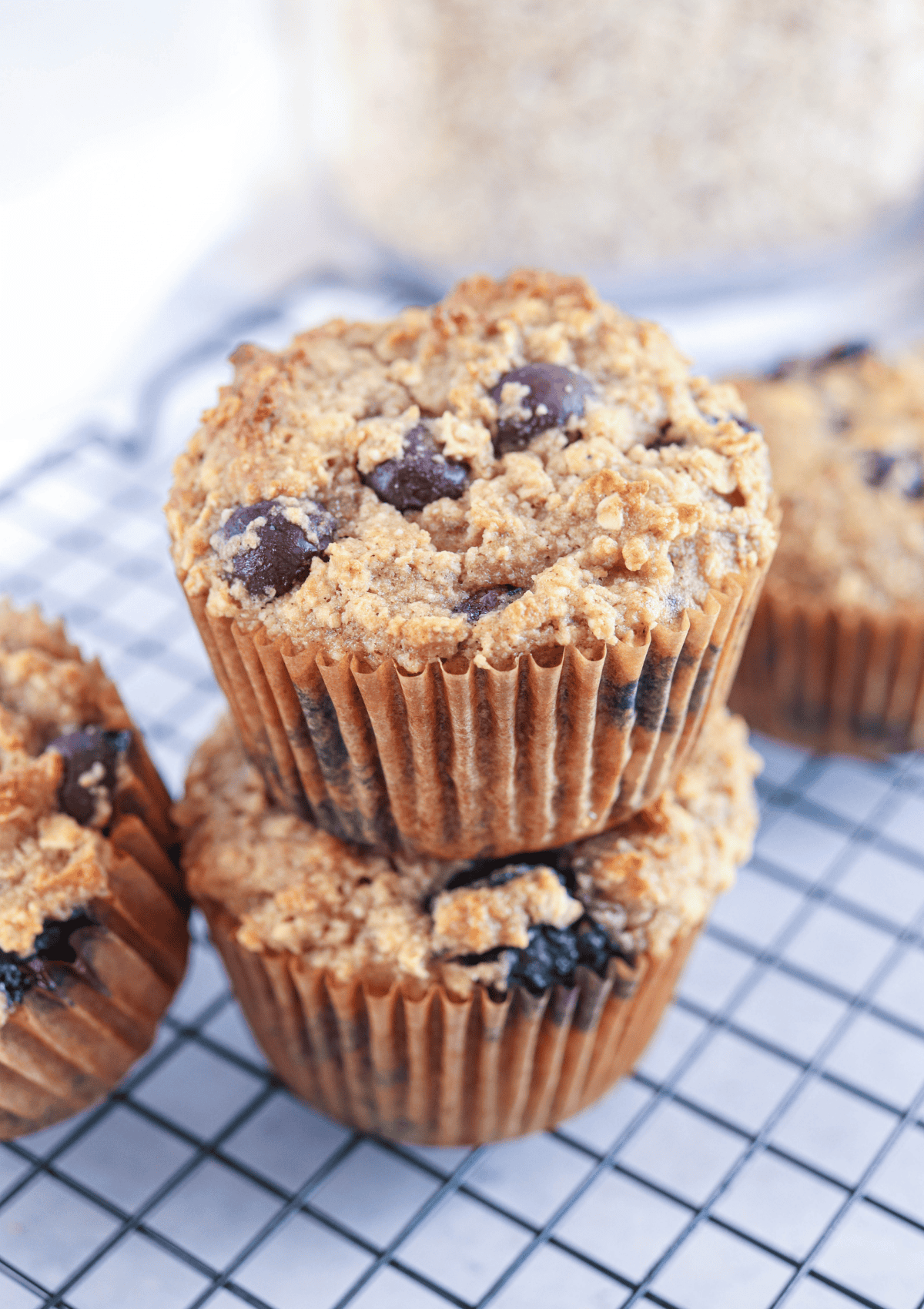 Three blueberry muffins in brown paper liners are stacked on a cooling rack, showcasing a cozy setup perfect for those seeking healthy banana breakfast recipes, with more muffins and a blurred container of oats in the background.