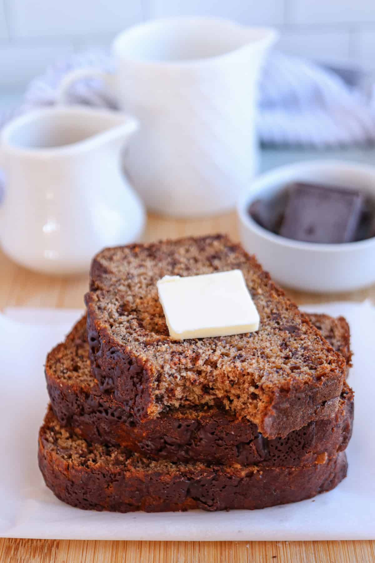 Three slices of banana bread stacked with a pat of butter on top, set on parchment paper with a jug, pitcher, and a bowl of chocolate piecesโperfect inspiration for healthy banana breakfast recipes.
