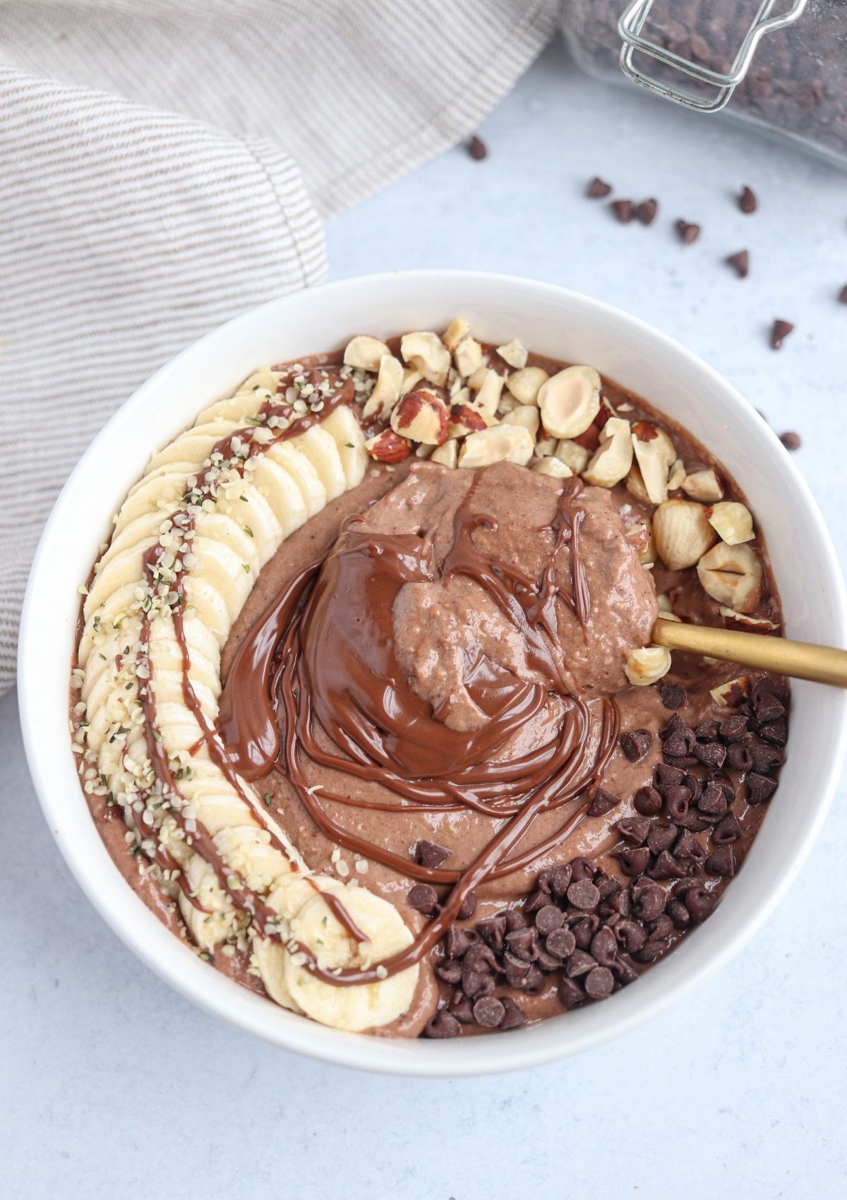 A white bowl filled with a chocolate smoothie topped with sliced banana, chopped hazelnuts, chocolate chips, hemp seeds, and a swirl of melted chocolateโone of the ultimate healthy banana breakfast recipesโwith a spoon inside.