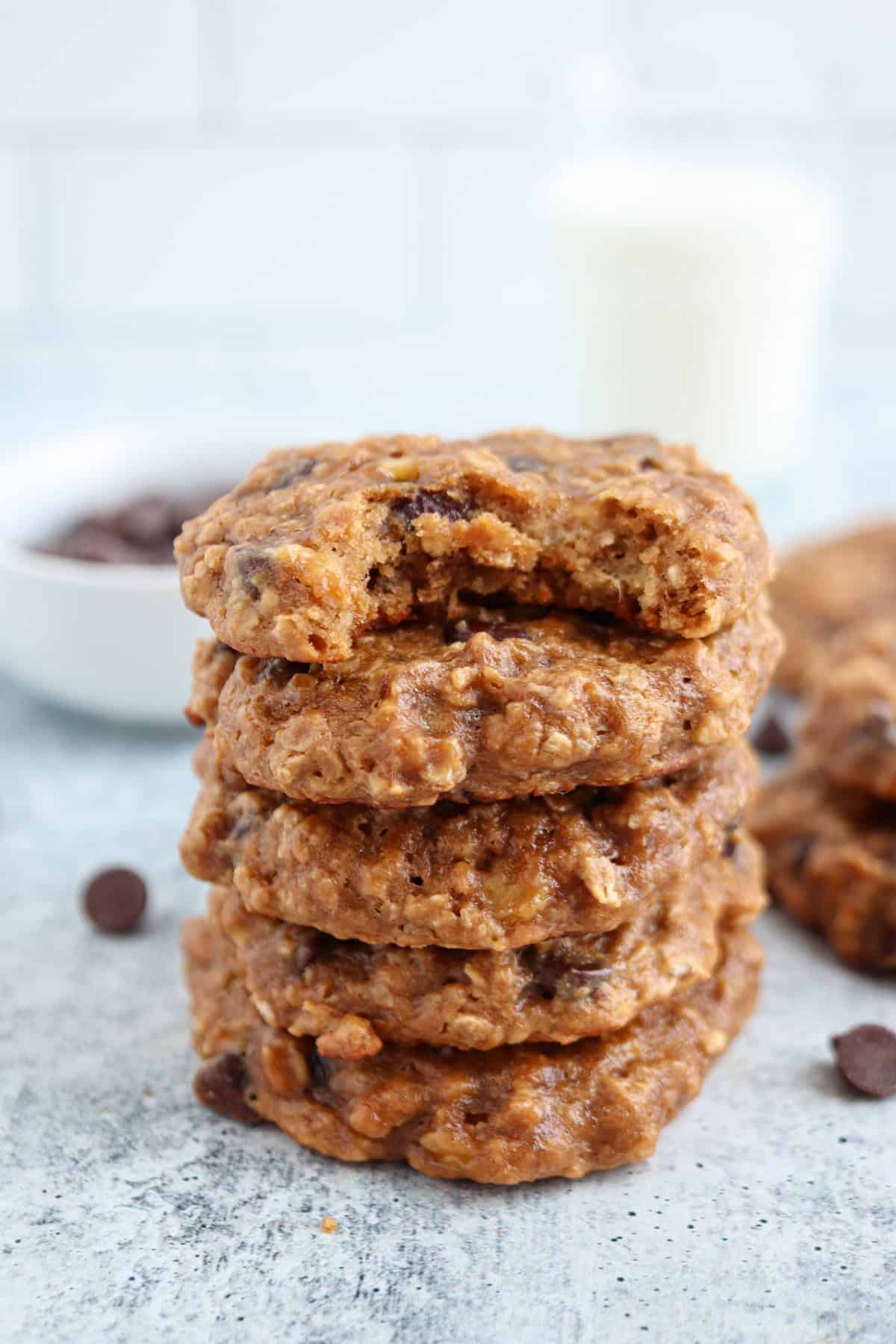 A stack of five oatmeal chocolate chip cookies, with the top cookie missing a bite. Perfect for healthy banana breakfast recipes, a bowl of chocolate chips and a glass of milk are blurred in the background.