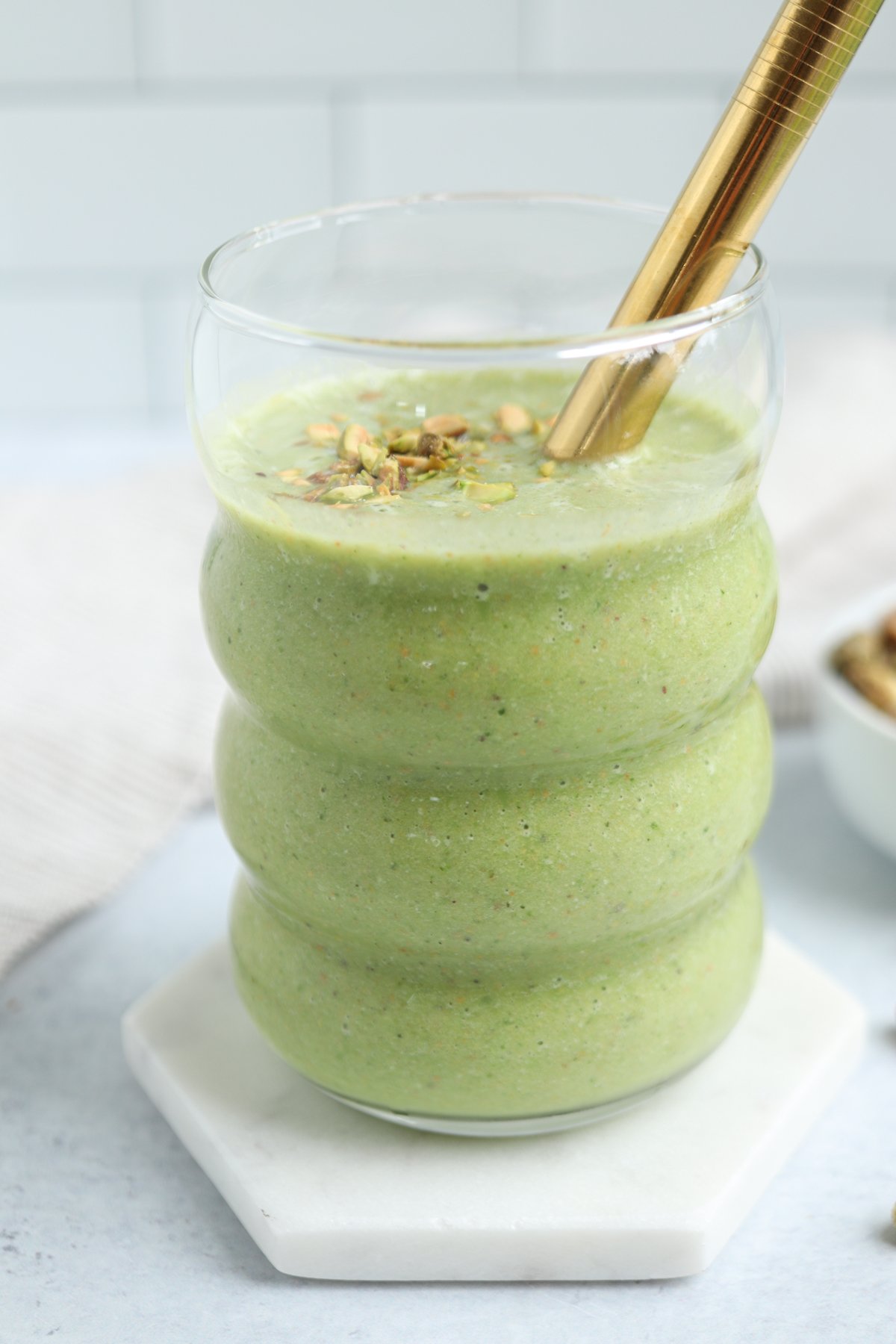 A thick green smoothie from healthy banana breakfast recipes is topped with chopped nuts, served in a wavy glass with a gold straw on a white hexagonal coaster. A small bowl of nuts sits in the background.